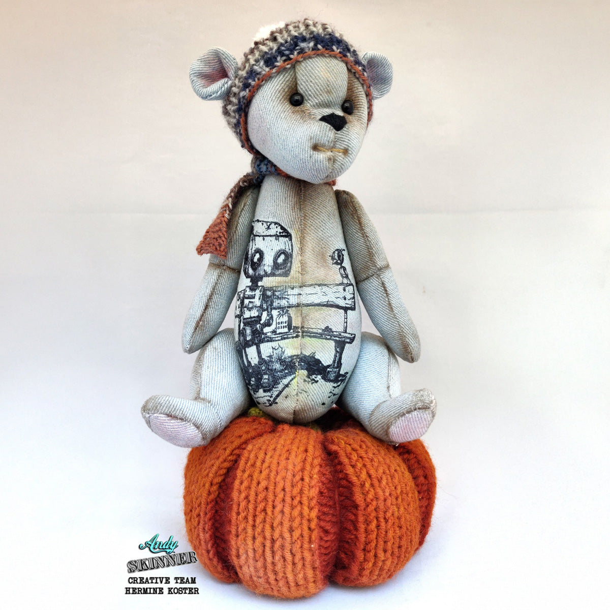 Teddy bear with spread a little kindness stamp design sitting on an orange knitted pumpkin base against a white background