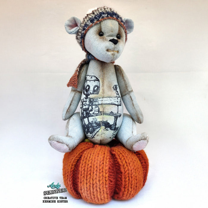 Teddy bear with spread a little kindness stamp design sitting on an orange knitted pumpkin base against a white background