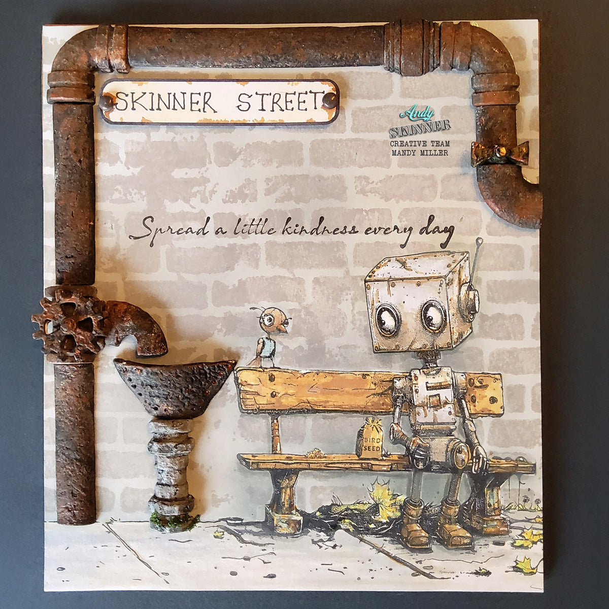 Robotic character sitting on a bench with 'Skinner Street' and 'Spread a little kindness every day' text on a brick wall background.